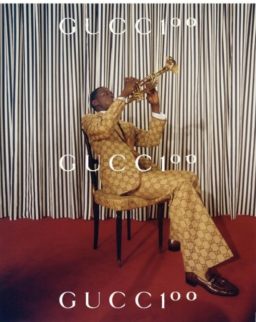 A man plays a trumpet while wearing a Gucci suit, seated in front of a striped backdrop, and the Gucci logo is visible in the scene.