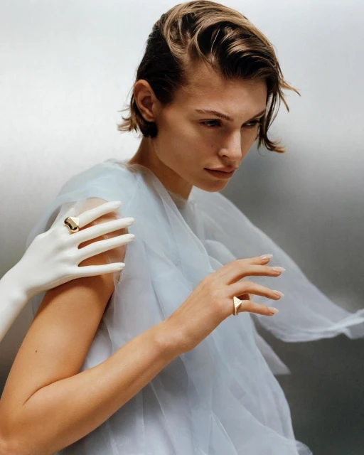 Portrait of a woman with short blonde hair wearing a flowing white dress and gold rings, posing with a mannequin hand in a studio.
