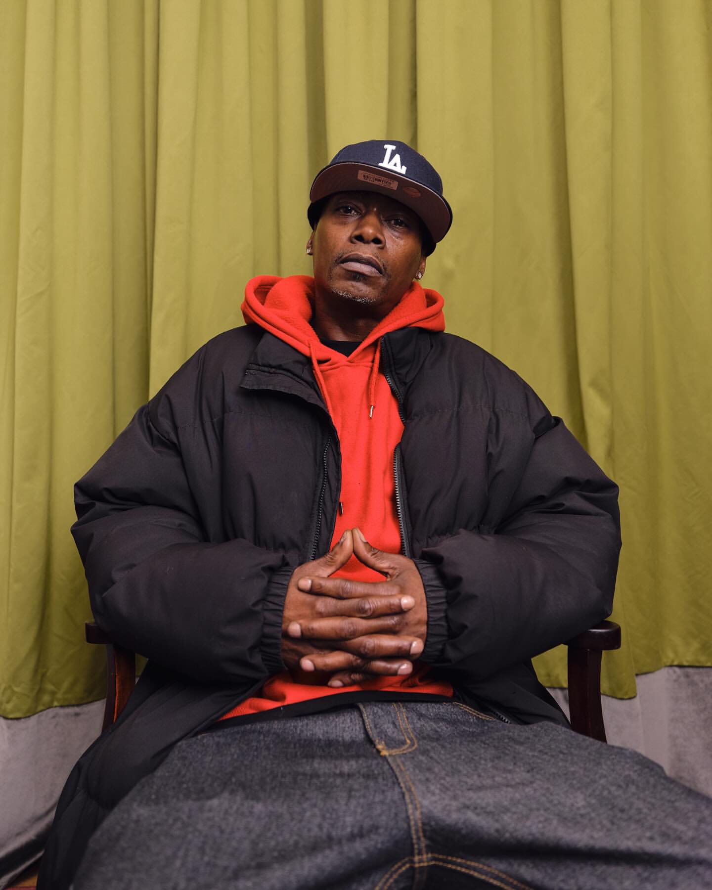 Black man wearing a black puffer jacket over a red hoodie and a baseball cap, seated with hands clasped.