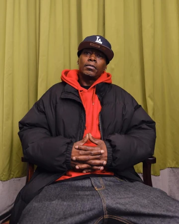 Black man wearing a black puffer jacket over a red hoodie and a baseball cap, seated with hands clasped.