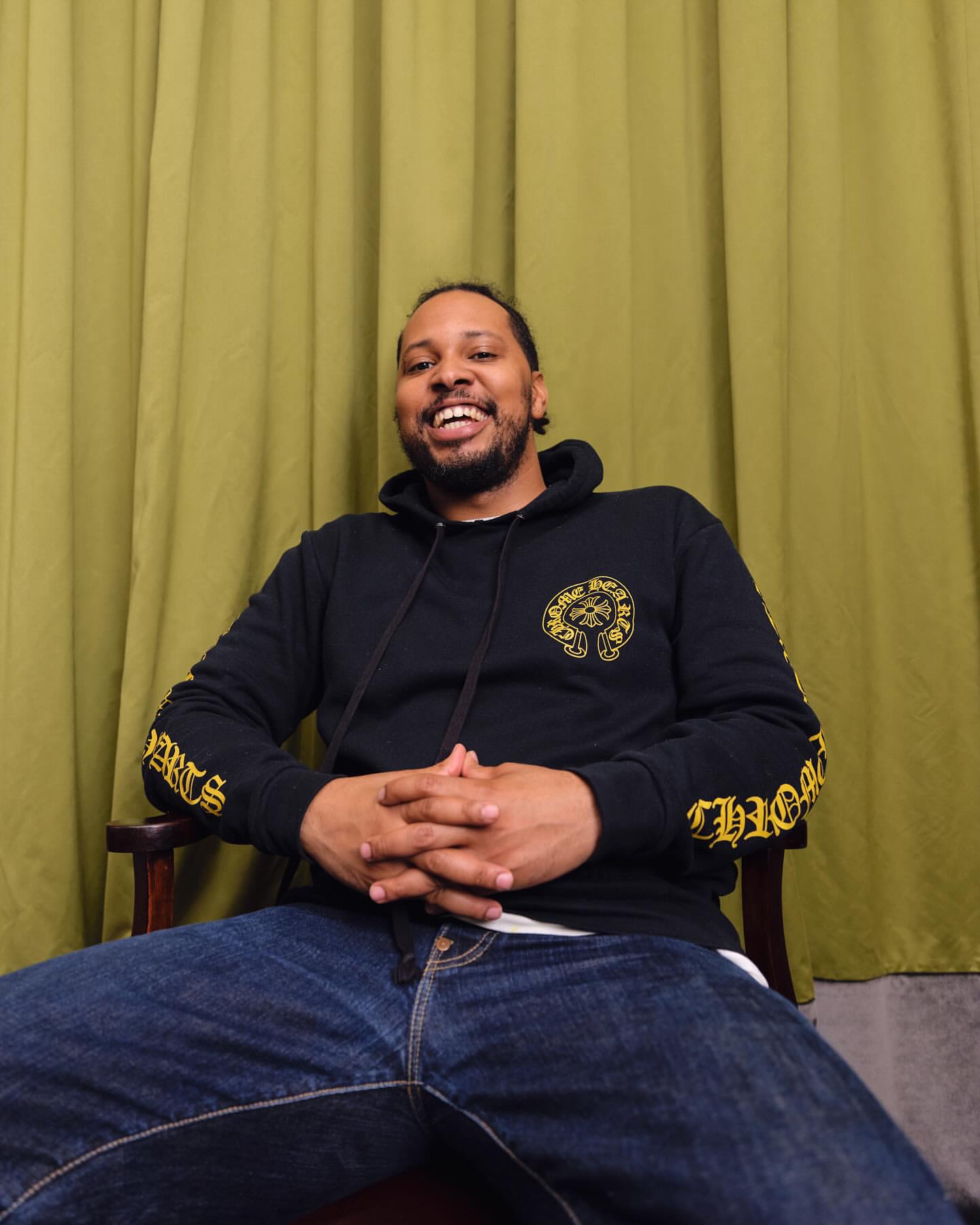 Portrait of a smiling Black man wearing a black hoodie and blue jeans, seated against an olive green curtain backdrop.