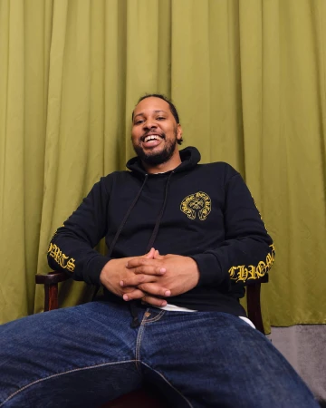 Portrait of a smiling Black man wearing a black hoodie and blue jeans, seated against an olive green curtain backdrop.