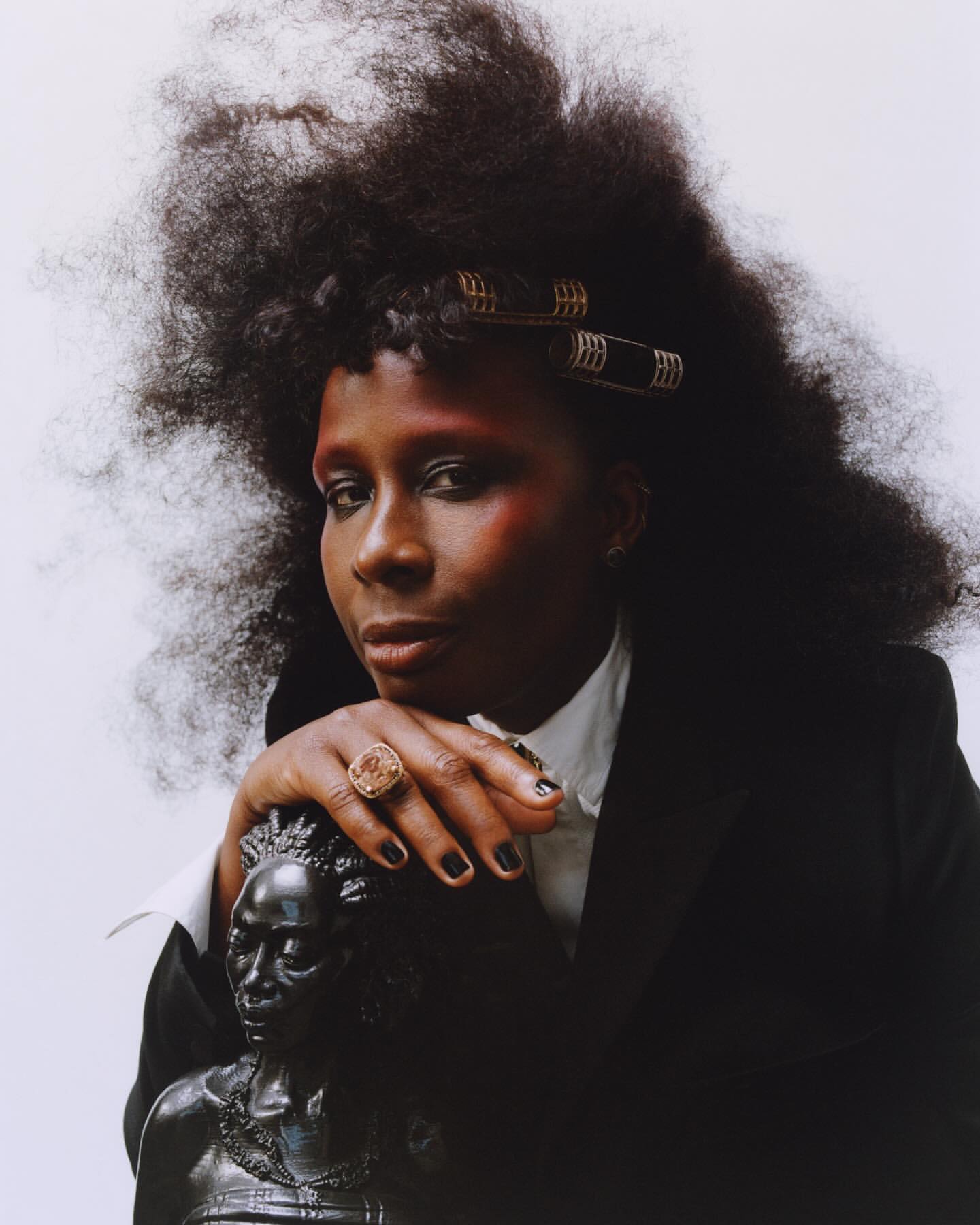 Portrait of an African-descent adult woman with large natural hair in curlers, wearing a black blazer, posing with a bust sculpture.