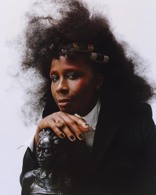 Portrait of an African-descent adult woman with large natural hair in curlers, wearing a black blazer, posing with a bust sculpture.