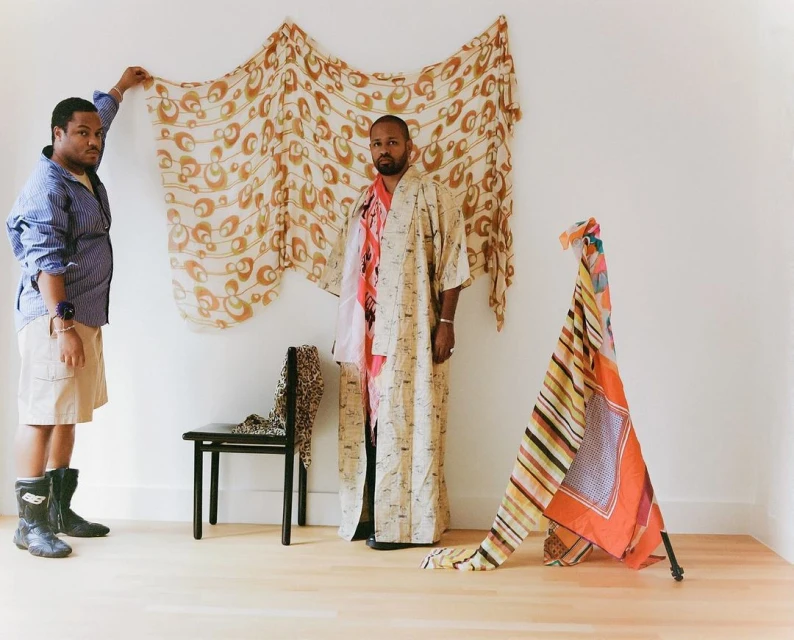 Two adult males in an indoor studio with light-colored walls, showcasing fabric art. One man holds fabric against the wall.
