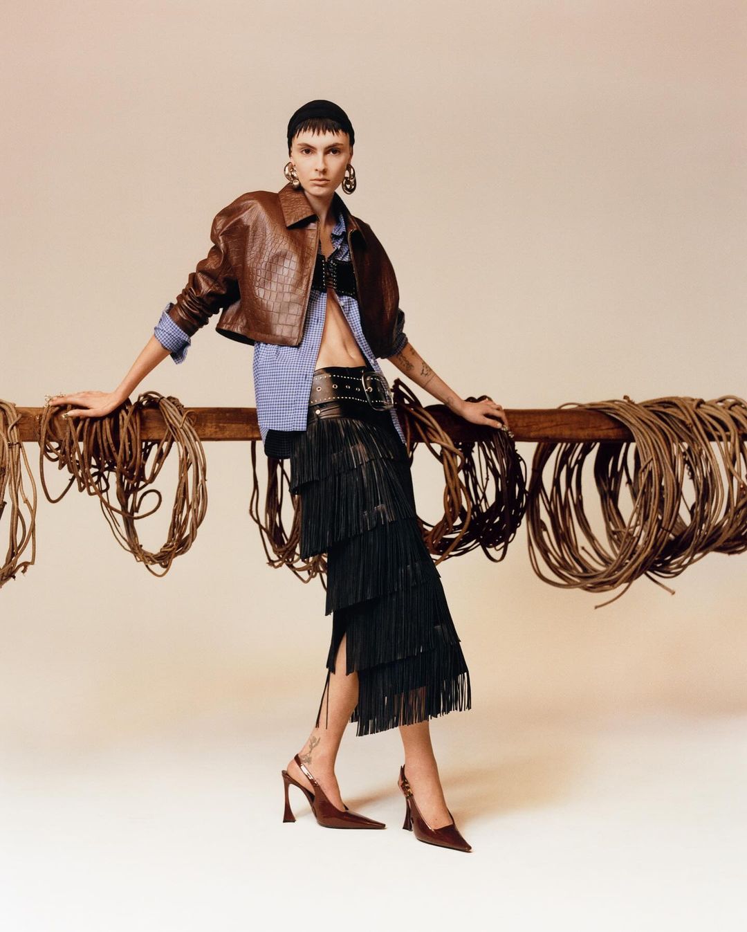 A woman poses in a brown leather jacket, striped shirt, black fringe skirt, and heels, leaning against a wooden rail with coiled ropes.