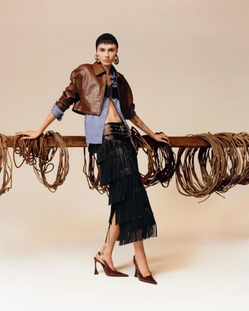 A woman poses in a brown leather jacket, striped shirt, black fringe skirt, and heels, leaning against a wooden rail with coiled ropes.
