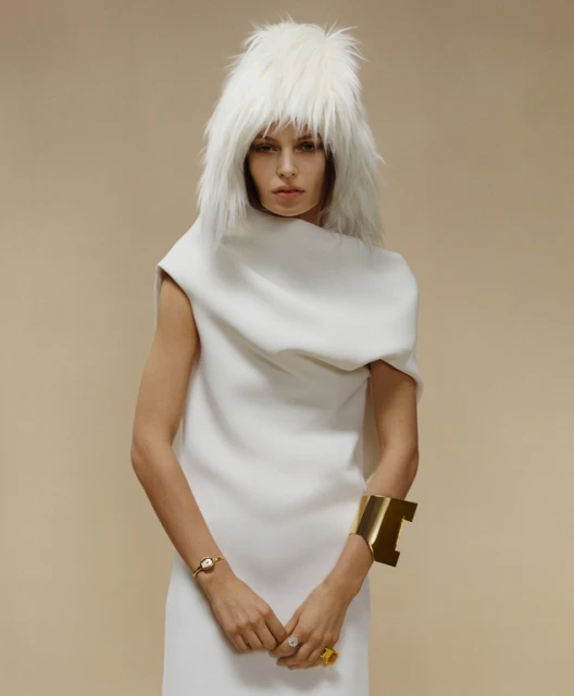 Elegant woman in a white dress and furry white hat poses against a neutral backdrop. Gold jewelry accents her wrists and fingers.