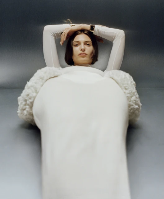 Portrait of a young woman with short brown hair lying on a silvery surface, wearing a long-sleeved white dress, a gold bracelet and a watch.