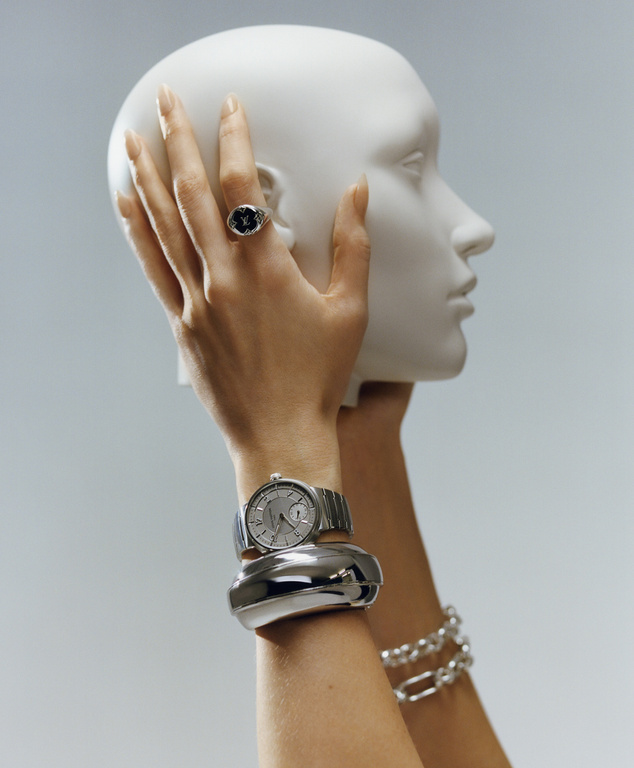 Close-up of a woman's arm with a silver watch and bracelets, holding a white mannequin head against a light blue backdrop.