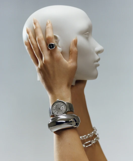 Close-up of a woman's arm with a silver watch and bracelets, holding a white mannequin head against a light blue backdrop.