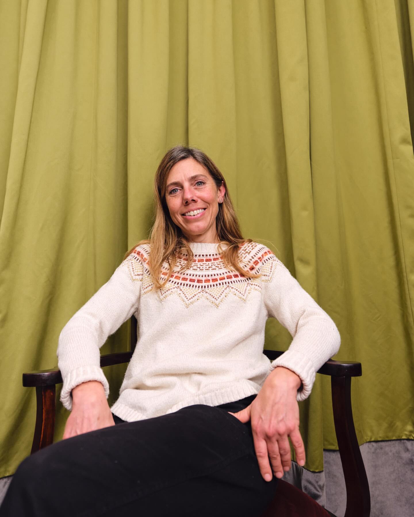 Woman sits in a wooden armchair against a solid green backdrop, wearing a cream-colored sweater with patterned yoke and a dark skirt.