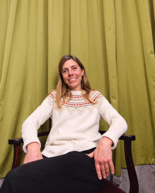 Woman sits in a wooden armchair against a solid green backdrop, wearing a cream-colored sweater with patterned yoke and a dark skirt.