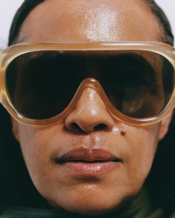 Close-up portrait of a person wearing translucent oversized sunglasses and glossy lip gloss photographed by Joshua Woods.