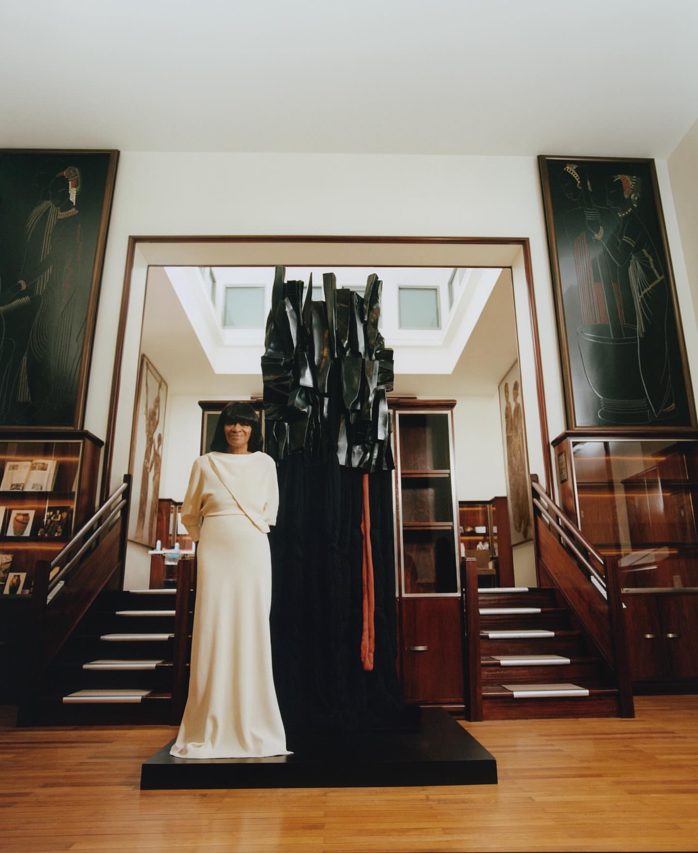 Woman in a long white dress standing next to a large dark sculptural installation in a wood-paneled room with stairs and framed artwork.