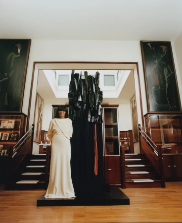 Woman in a long white dress standing next to a large dark sculptural installation in a wood-paneled room with stairs and framed artwork.