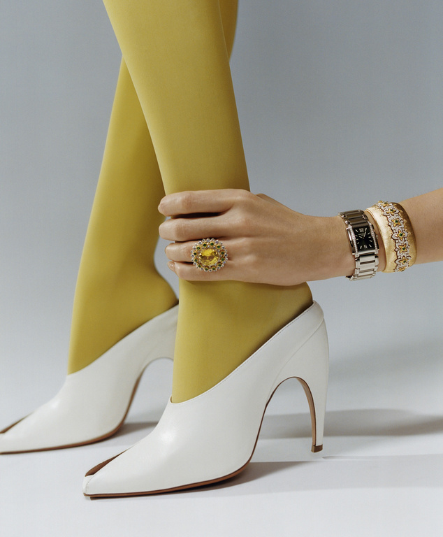 Close-up of woman's legs in yellow stockings and white heels, adorned with jewelry on hand, wrist, and finger on a light grey background.