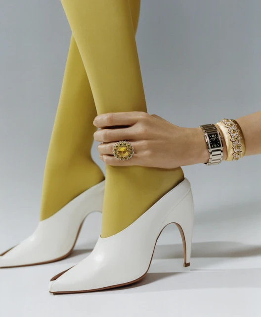 Close-up of woman's legs in yellow stockings and white heels, adorned with jewelry on hand, wrist, and finger on a light grey background.