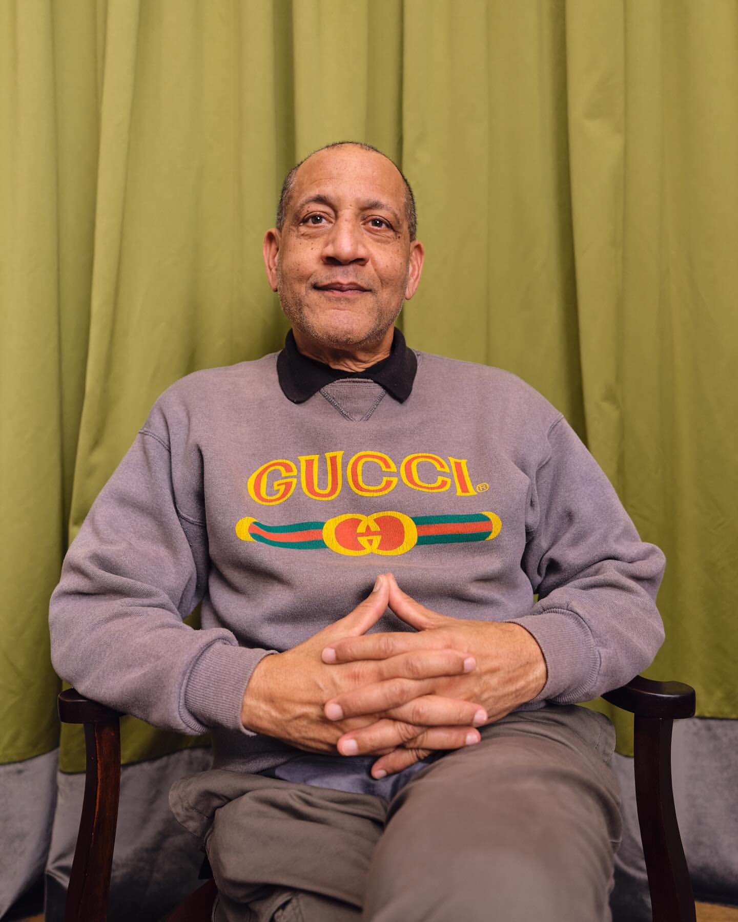 An African American man in a Gucci sweatshirt sits in a chair against an olive-green curtain backdrop, hands clasped, in a medium shot.