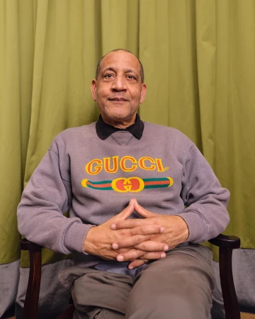 An African American man in a Gucci sweatshirt sits in a chair against an olive-green curtain backdrop, hands clasped, in a medium shot.