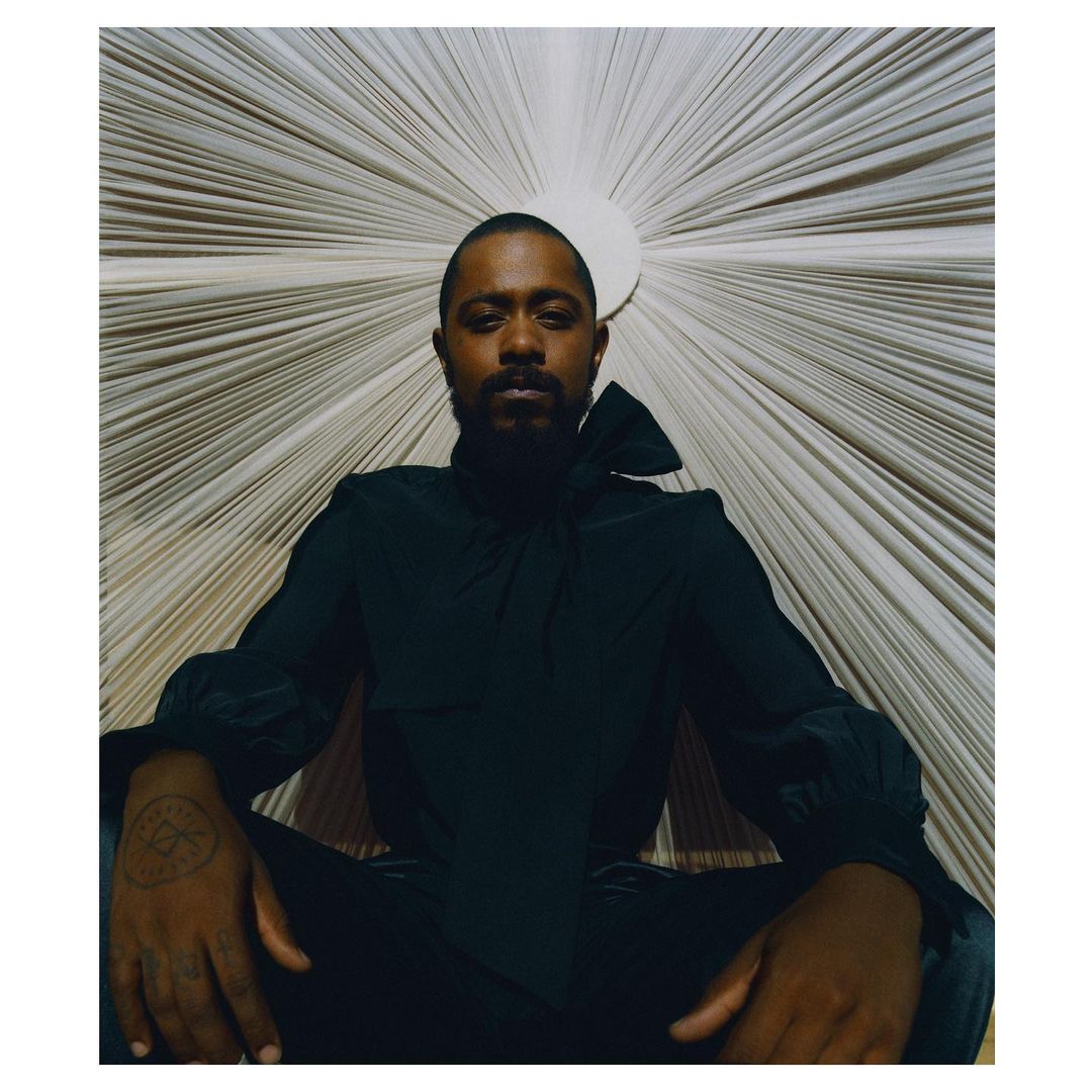 Portrait of a Black adult male with a beard, wearing a black top and pants, sitting in front of a neutral sunburst-patterned background.