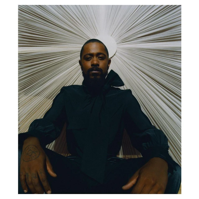 Portrait of a Black adult male with a beard, wearing a black top and pants, sitting in front of a neutral sunburst-patterned background.