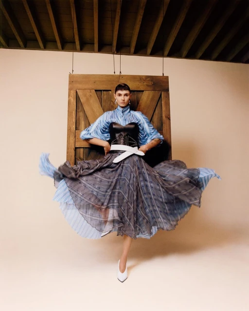 A stylish woman in a blue blouse, black corset, and flowing skirt poses in front of a wooden door with neutral studio background.