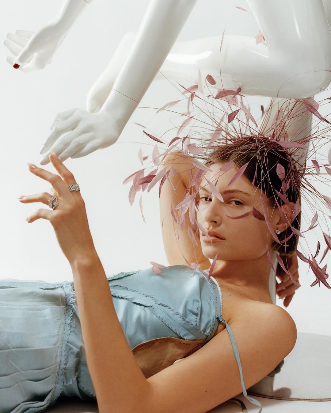 Woman poses with a feather headpiece and a light blue corset dress next to a mannequin hand in a brightly lit indoor photo studio.