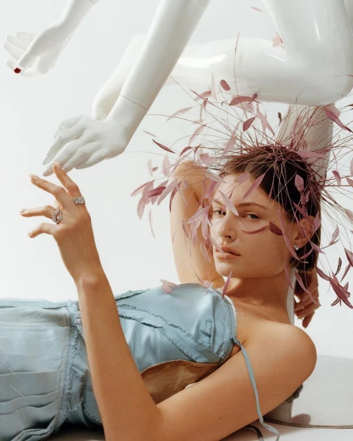 Woman poses with a feather headpiece and a light blue corset dress next to a mannequin hand in a brightly lit indoor photo studio.