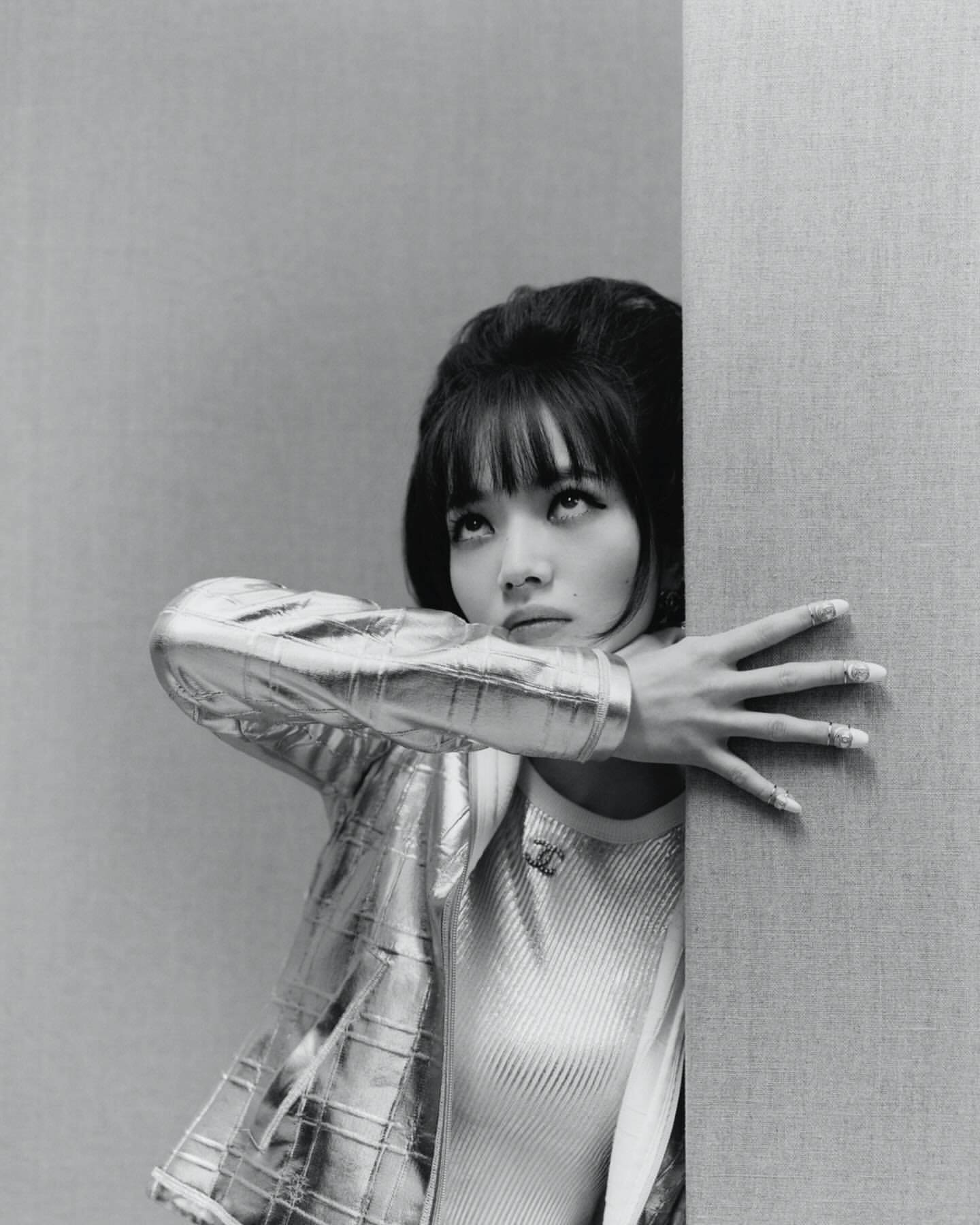 Stylish woman with bangs, wearing a metallic jacket, leans against a wall in a close-up studio portrait with monochrome tones.