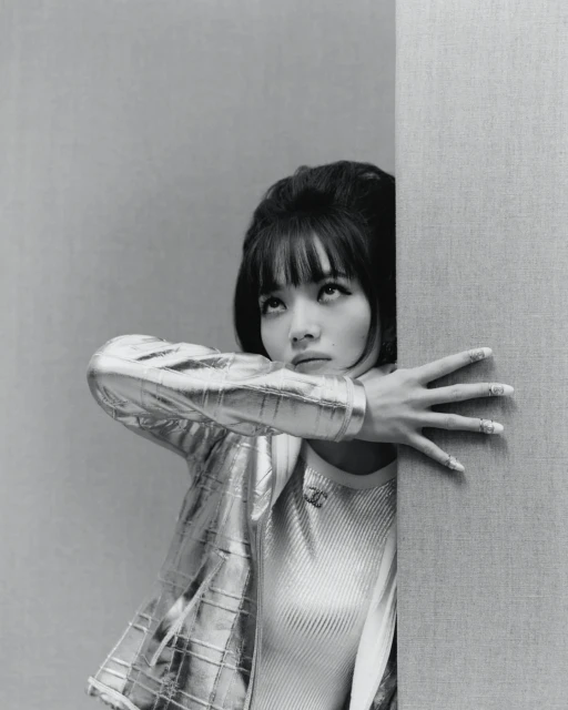 Stylish woman with bangs, wearing a metallic jacket, leans against a wall in a close-up studio portrait with monochrome tones.