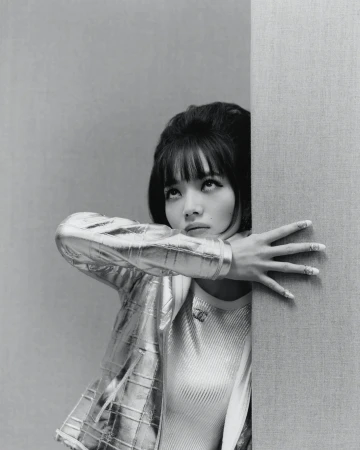 Stylish woman with bangs, wearing a metallic jacket, leans against a wall in a close-up studio portrait with monochrome tones.
