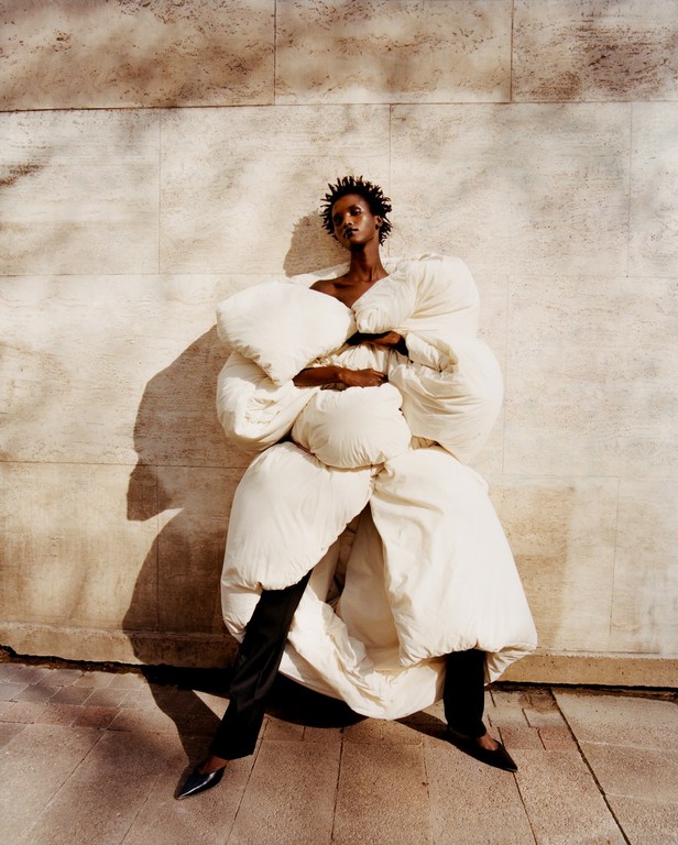 Fashionable black model in an oversized white down coat poses against a textured beige wall in an outdoor fashion photography shot.