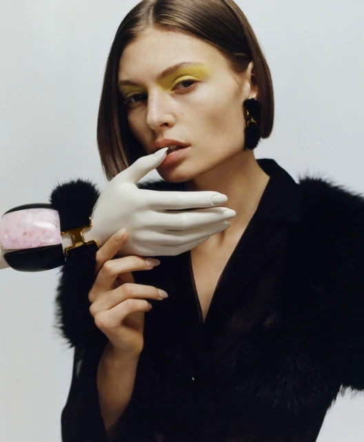 Close-up of a fair-skinned woman with yellow eyeshadow and bob haircut, wearing a black top with a prosthetic hand on her neck.