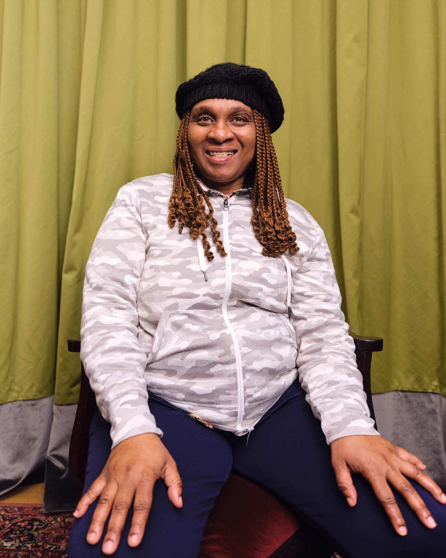 Portrait of a mature Black woman in a black beret and grey patterned zip-up jacket, smiling indoors with a green curtain backdrop.