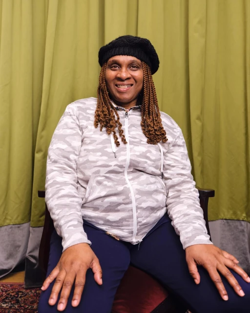 Portrait of a mature Black woman in a black beret and grey patterned zip-up jacket, smiling indoors with a green curtain backdrop.