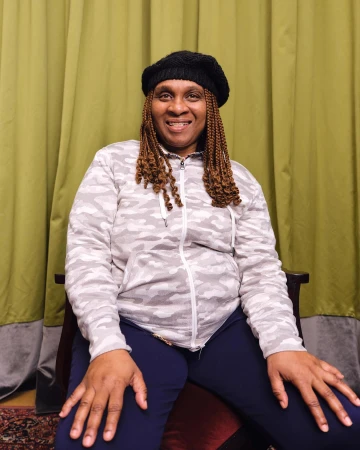 Portrait of a mature Black woman in a black beret and grey patterned zip-up jacket, smiling indoors with a green curtain backdrop.