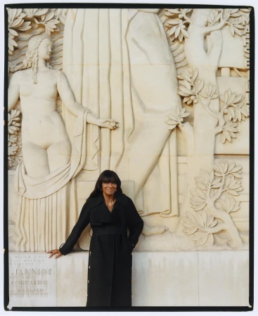 Adult female poses next to a Beaux-Arts style architectural stone carving featuring a nude female figure and a tree motif.