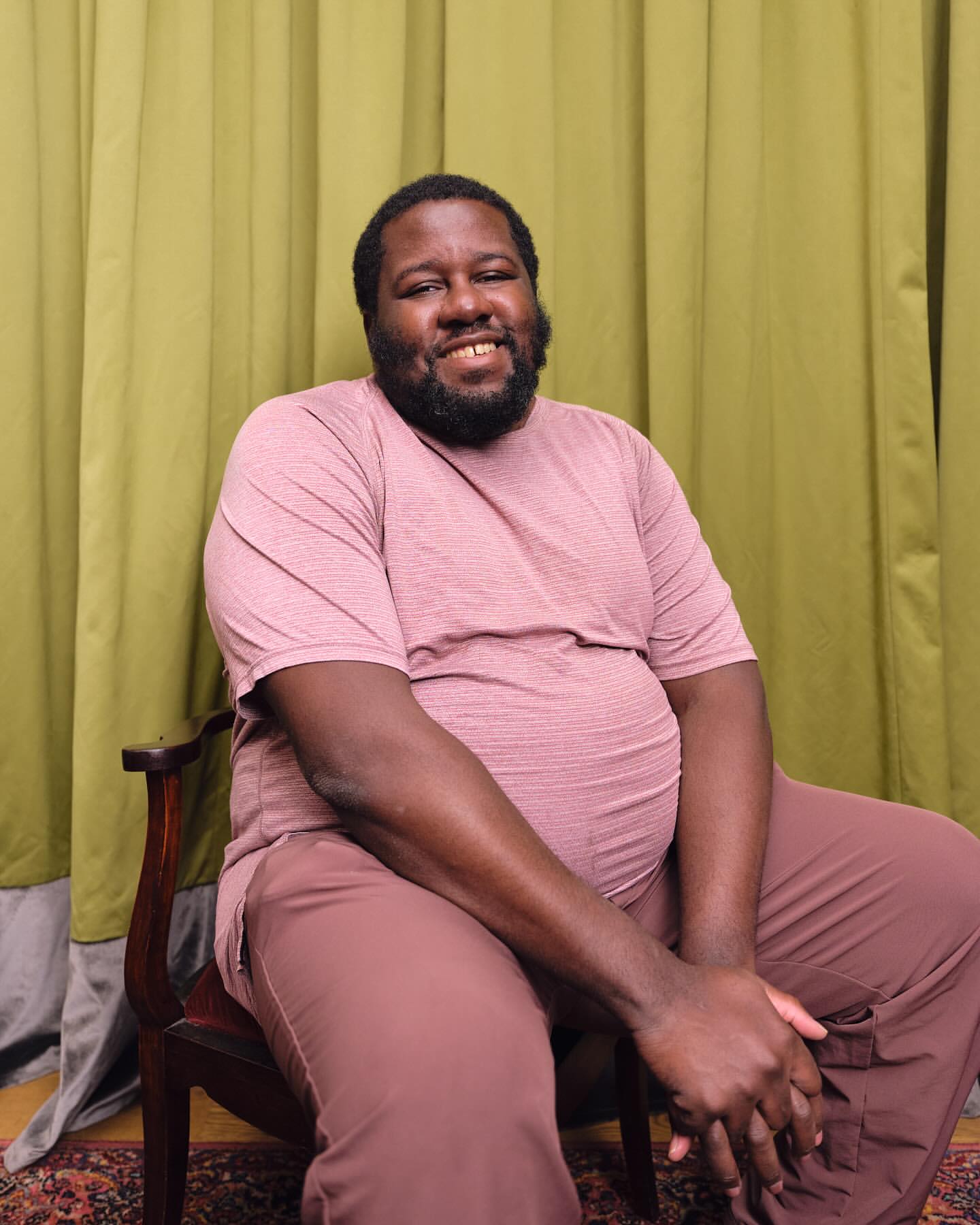 Portrait of a smiling adult male with dark hair and a beard seated on a chair with a green curtain backdrop in an indoor setting.