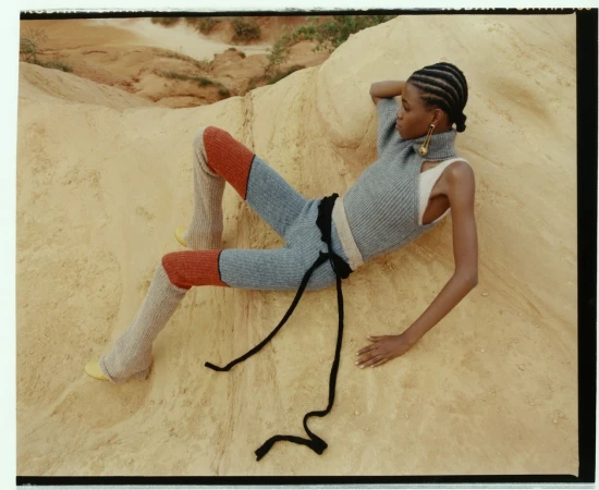 Black model with braided hair wearing grey knit apparel and orange leggings reclining on sandy terrain