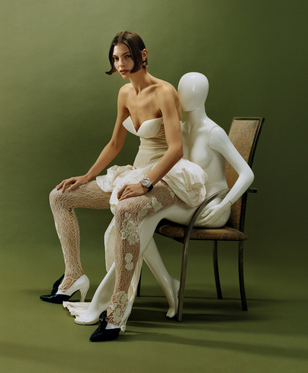 Fashion shot of a woman in a dress sitting on a chair with a mannequin, in front of a green background. The woman is wearing heels.