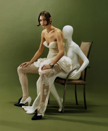 Woman in strapless white dress and lace tights seated on a white mannequin against an olive green backdrop
