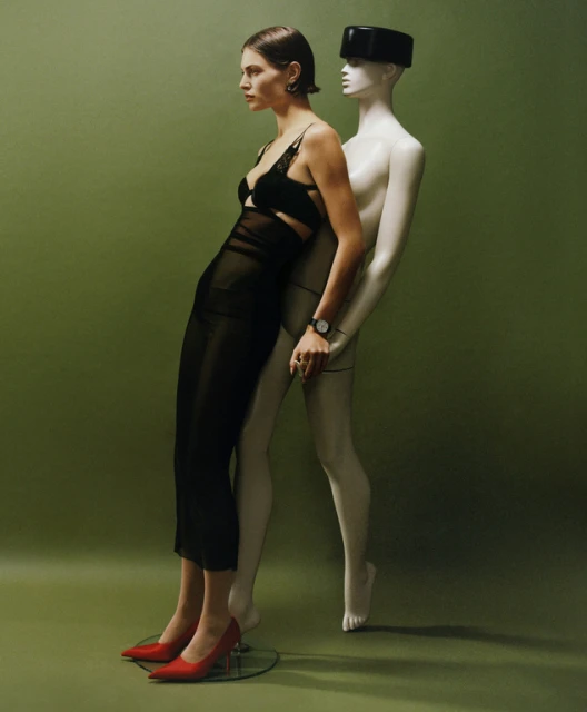 A woman in a black dress and red heels leans against a mannequin in a studio setting with a light green background and soft lighting.