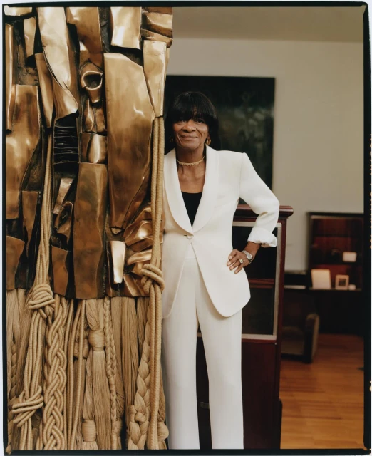 Elegant woman in a white suit poses next to a gold-toned abstract sculpture in an artful indoor setting with neutral walls and wood accents.
