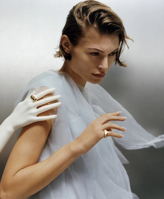 Elegant portrait of a woman with a mannequin hand adorned with a ring, set against a soft grey backdrop with shallow depth of field.