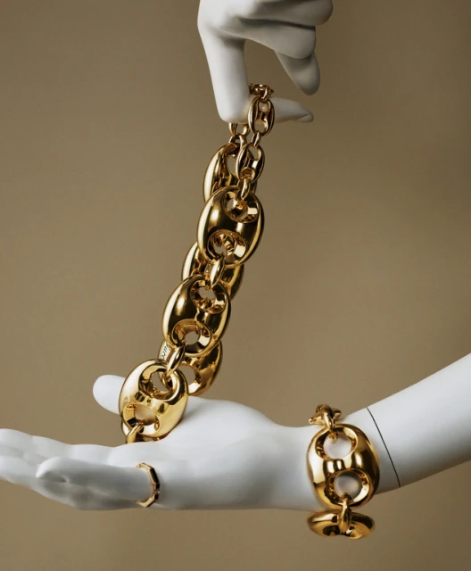 Gold chain link jewelry is presented on white mannequin hands against a neutral background in a clean studio setting. Fashion shot.
