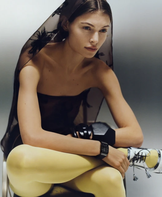 Portrait of a fair-skinned woman in sheer black headscarf, strapless top, yellow leggings, wearing a black watch, seated indoors.