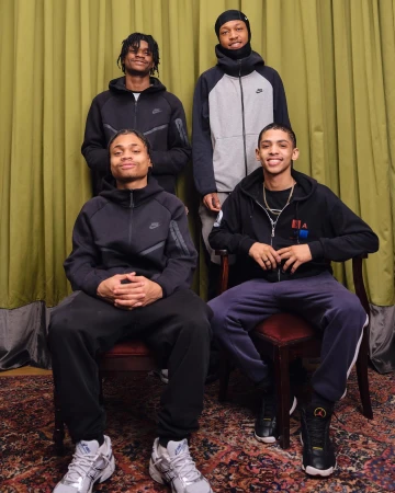 A group of four young adult males in sportswear posing indoors against a green curtain backdrop, seated and standing, medium shot.