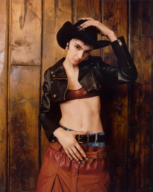 Woman in cowboy hat, leather jacket and skirt poses against a wood wall, looking at the camera, in a fashion editorial style.