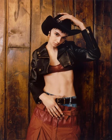 Woman in cowboy hat, leather jacket and skirt poses against a wood wall, looking at the camera, in a fashion editorial style.